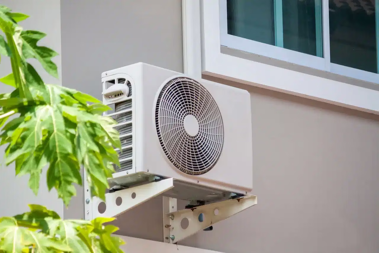 Outdoor air conditioner compressor mounted beside a home with green plants