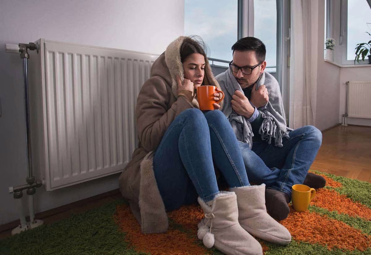 Couple in coats huddled by a radiator with mugs in a cold home